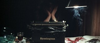 Una habitación oscura con un escritorio sobre el que hay una máquina de escribir negra de la marca Remington. El escritorio está cubierto de libros abiertos, papeles y un dibujo con líneas rojas. A la izquierda hay una copa con una bebida oscura y un objeto de vidrio azul. A la derecha cuelga una lámpara, bajo la cual se eleva humo. Detrás de la máquina de escribir está sentada una persona, con la cabeza apoyada en las manos.