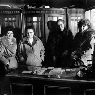 Interior de una tienda antigua con un mostrador de madera en primer plano. Detrás del mostrador hay varias mujeres con abrigos de invierno. Al fondo se observan armarios altos de madera. A la derecha aparece una caja registradora histórica con un cartel que dice “nachzuzählen”.
