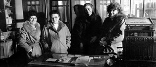 Interior de una tienda antigua con un mostrador de madera en primer plano. Detrás del mostrador hay varias mujeres con abrigos de invierno. Al fondo se observan armarios altos de madera. A la derecha aparece una caja registradora histórica con un cartel que dice “nachzuzählen”.
