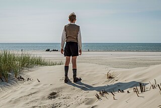 Eine Person steht auf einem sandigen Strand und blickt auf das Meer hinaus. Sie befindet sich in der Mitte des Bildes, trägt einen langärmligen Pullover, einen kurzen Rock und Stiefel. Im Vordergrund liegen helle Sanddünen mit vereinzelten Grasbüscheln. Hinter der Person erstreckt sich das ruhige Meer bis zum Horizont, darüber ein klarer blauer Himmel. 