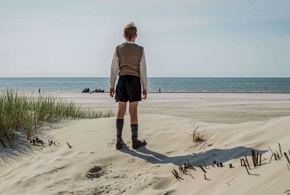 Eine Person steht auf einem sandigen Strand und blickt auf das Meer hinaus. Sie befindet sich in der Mitte des Bildes, trägt einen langärmligen Pullover, einen kurzen Rock und Stiefel. Im Vordergrund liegen helle Sanddünen mit vereinzelten Grasbüscheln. Hinter der Person erstreckt sich das ruhige Meer bis zum Horizont, darüber ein klarer blauer Himmel. 