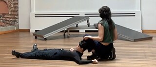 En primer plano se ve a dos personas bailando. Una persona está tumbada boca arriba y tiene la cabeza ligeramente apoyada en la pierna de la otra persona, que está sentada a su derecha, de espaldas al público.