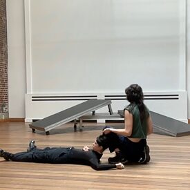 Zwei tanzende Personen sind im Vordergrund gesehen. Eine Person liegt auf dem Rücken und hat den Kopf leicht am Bein der anderen Person angelehnt, die rechts neben ihm vom Publikum weggedreht hockt.