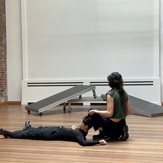 Zwei tanzende Personen sind im Vordergrund gesehen. Eine Person liegt auf dem Rücken und hat den Kopf leicht am Bein der anderen Person angelehnt, die rechts neben ihm vom Publikum weggedreht hockt.