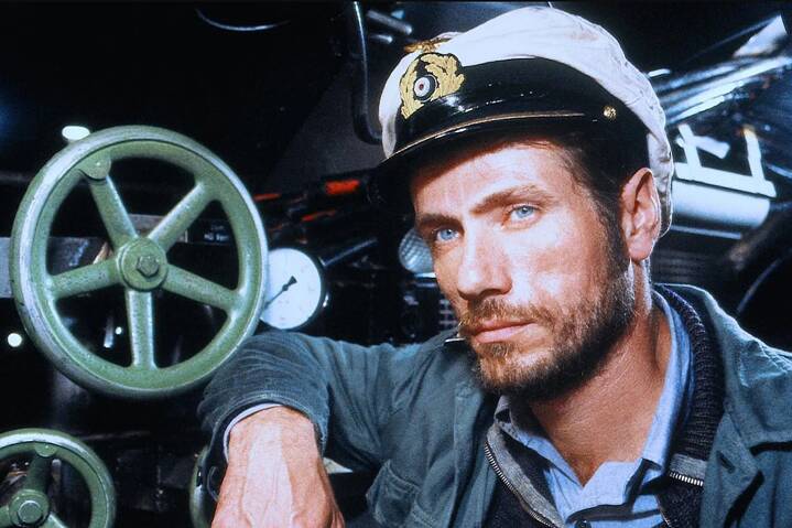 Standbild aus "Das Boot". Ein Mann in einer Marineuniform mit Offiziersmütze steht vor einem technischen Bedienpult. Hinter ihm befinden sich große grüne Metallräder und mehrere Rundinstrumente. Sein Gesicht ist ernst, der Raum ist eng und von gedämpftem Licht geprägt. Die Umgebung wirkt wie das Innere eines U‑Boots mit sichtbaren mechanischen Elementen.