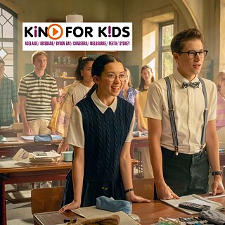 Eine Klassenzimmerszene mit mehreren Schülern, die zwischen Holztischen voller Bücher, Papiere und Schulmaterialien stehen oder sitzen.