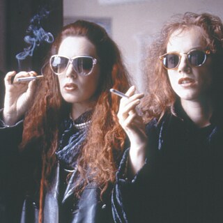 Dos mujeres con cabello largo y rizado están en un espacio interior y sostienen cigarrillos encendidos. Llevan ropa oscura y se ve humo elevándose en el aire.