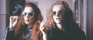 Dos mujeres con cabello largo y rizado están en un espacio interior y sostienen cigarrillos encendidos. Llevan ropa oscura y se ve humo elevándose en el aire.