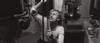 Still aus dem Film "Im Lauf der Zeit" von Wim Wenders: Rüdiger Vogler sitzt in einem kleinen, mit Filmtechnik ausgestatteten Arbeitsraum und hält einen längeren Streifen analoges Filmmaterial senkrecht nach oben, um ihn zu prüfen oder zu bearbeiten.