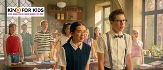 Eine Klassenzimmerszene mit mehreren Schülern, die zwischen Holztischen voller Bücher, Papiere und Schulmaterialien stehen oder sitzen.