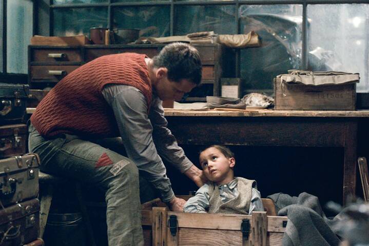Ein Mann in einem roten Strickwesten beugt sich über einen kleinen Jungen und hebt ihn vorsichtig in eine hölzerne Kiste. Der Junge trägt ein helles Hemd und eine graue Weste und blickt ruhig nach oben. Die Umgebung ist ein dunkler Raum mit alten Holzmöbeln, Koffern und Kisten, die dicht beieinander stehen. Das Licht ist gedämpft und fällt von der Seite auf die beiden Figuren.