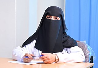 امرأة تجلس إلى طاولة ممسكةً بقلم. ترتدي نقابًا أسود وتنظر مباشرةً إلى الكاميرا.