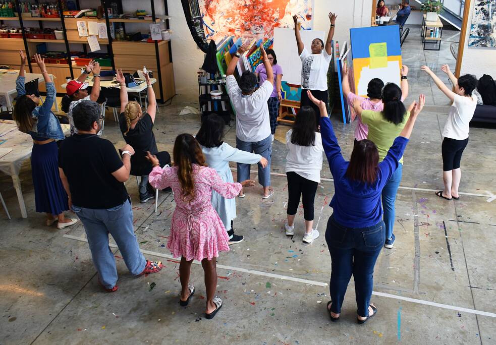 Men and women are standing in a room raising their arms. In the background you can see paintings and art supplies. 