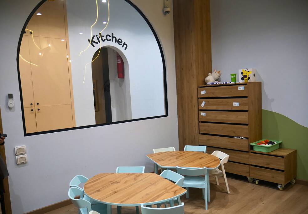 فصل دراسي بطاولات وكراسي صغيرة. رف كتب. من النافذة يُمكن رؤية باب المطبخ.