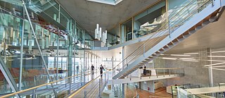 Ein großer, moderner Innenraum mit mehreren Ebenen, Glaswänden und offenen Treppenanlagen. Ein weitläufiges Netz aus Metallgeländern, Holzstegen und schwebenden Treppen verbindet verschiedene Stockwerke. Im Zentrum verlaufen breite Treppen und Rampen mit Holzbelag. Auf der linken Seite des Bildes spiegeln große Glasflächen den Innenraum und zeigen dahinterliegende Arbeits- oder Aufenthaltsbereiche mit Tischen und Stühlen. An der Decke hängen markante, vertikal angeordnete Leuchten. Im oberen Bereich fallen Tageslichtöffnungen und große Fenster ins Gebäude. Einige Personen bewegen sich auf den Wegen und Treppen durch den Raum.