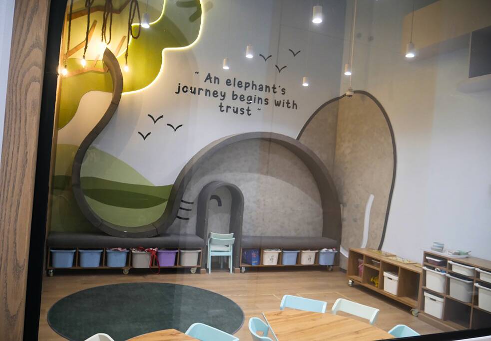 فصل دراسي. على الجدار رُسم رأس فيل، وشجرة، وطيور نورس بسيطة. كُتب على الجدار: "رحلة الفيل تبدأ بالثقة".