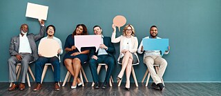 Des hommes et des femmes en tenue d'affaires, assis en ligne les uns à côté des autres, tiennent de grandes bulles colorées devant eux ou en l'air. Ils s'amusent et rient face à la caméra. Espace pour le texte rédactionnel.