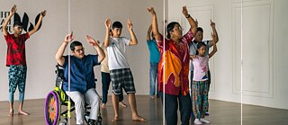 Adults and children are practicing dance in a dance studio infront of a mirror. One man is sitting in a wheelchair. 