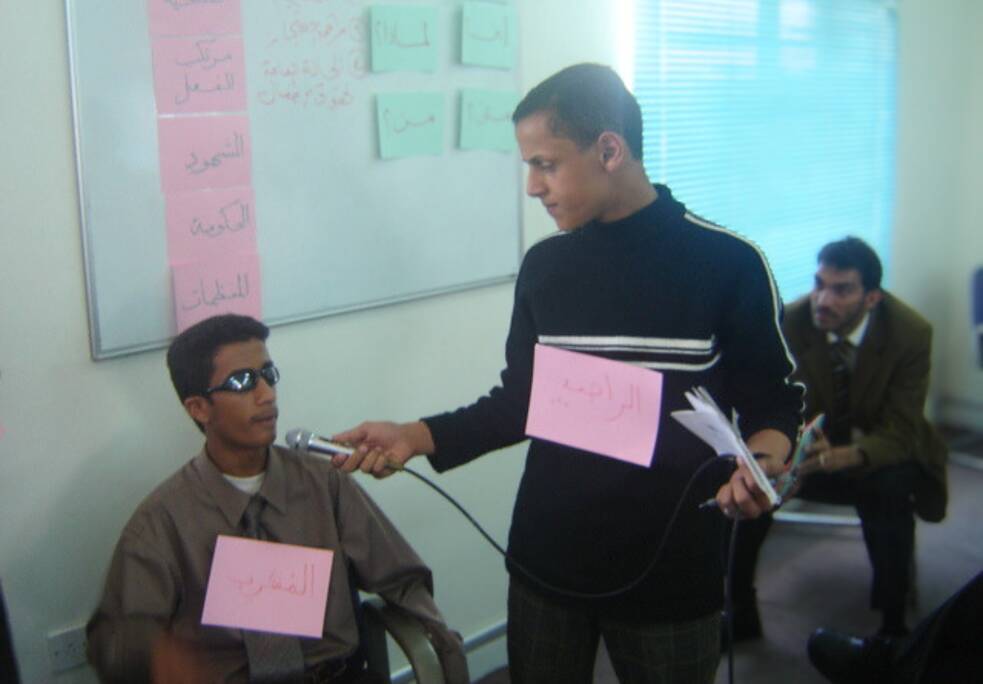 يجلس فتى مراهق على كرسي في قاعة دراسية، يرتدي نظارة شمسية داكنة اللون، ويعلق لافتة حول عنقه. يقف بجانبه شاب آخر، يرتدي هو الآخر لافتة حول عنقه، ويحمل ميكروفونًا موجهًا نحو وجه الفتى المراهق.