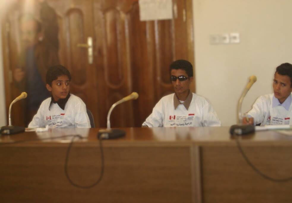 ثلاثة فتيان مراهقين يجلسون حول طاولة اجتماعات مزودة بميكروفونات.