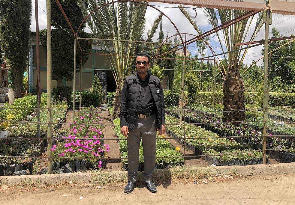 رجل يقف أمام حديقة. يرتدي نظارة شمسية ويبتسم للكاميرا.