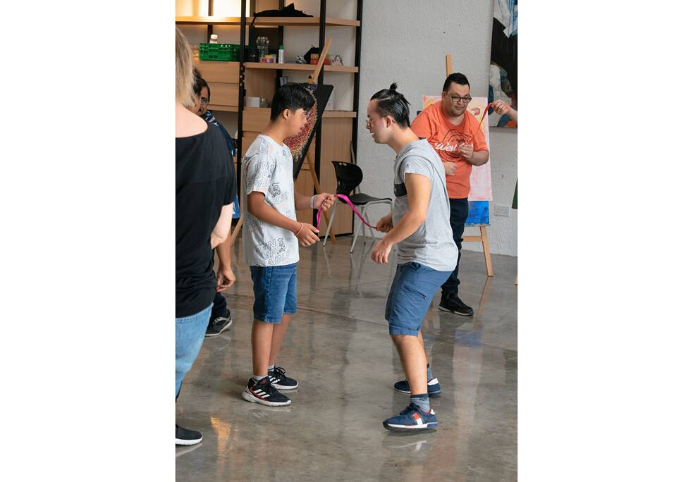 Two teenage boys stand opposite to eachother and hold a pink cord in one hand. Behind and in front of them are other teenager doing the same. 