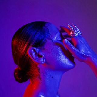 Nahaufnahme eines Porträts: Eine Person mit zu einem tiefen Dutt gebundenem Haar wird von intensiven blauen und roten Lichtfarben beleuchtet. Die Augen sind geschlossen, eine Hand berührt das Gesicht und zeigt einen auffälligen, organisch geformten Ring. Die Haut wirkt glänzend, fast feucht, was eine dramatische und ästhetische Stimmung erzeugt.