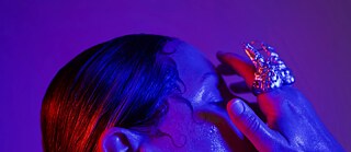 Nahaufnahme eines Porträts: Eine Person mit zu einem tiefen Dutt gebundenem Haar wird von intensiven blauen und roten Lichtfarben beleuchtet. Die Augen sind geschlossen, eine Hand berührt das Gesicht und zeigt einen auffälligen, organisch geformten Ring. Die Haut wirkt glänzend, fast feucht, was eine dramatische und ästhetische Stimmung erzeugt.