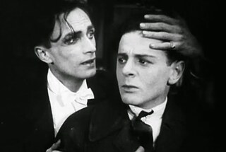 Conrad Veidt und Fritz Schulz in „Anders als die Anderen“: Das schwarz‑weiße Bild zeigt zwei Personen in Nahaufnahme. Die linke Person trägt einen dunklen Anzug mit weißem Hemd und weißer Fliege und legt eine Hand auf den Kopf der rechten Person. Die rechte Person blickt nach vorn und trägt ebenfalls formelle Kleidung. Der Hintergrund ist dunkel, und die Szene ist stark ausgeleuchtet, sodass die Gesichter deutlich sichtbar sind.