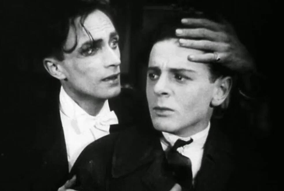 Conrad Veidt und Fritz Schulz in „Anders als die Anderen“: Das schwarz‑weiße Bild zeigt zwei Personen in Nahaufnahme. Die linke Person trägt einen dunklen Anzug mit weißem Hemd und weißer Fliege und legt eine Hand auf den Kopf der rechten Person. Die rechte Person blickt nach vorn und trägt ebenfalls formelle Kleidung. Der Hintergrund ist dunkel, und die Szene ist stark ausgeleuchtet, sodass die Gesichter deutlich sichtbar sind.