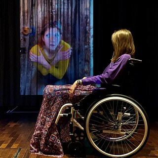 Ausschnitt aus „Die Gipsfabrik“: Die Schauspielerin sitzt im Rollstuhl auf der Bühne und betrachtet ihr eigenes Porträt, das auf die Leinwand hinter der Bühne projiziert wird.