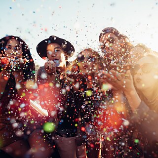 Gruppe von Personen im Freien, die dicht beieinandersteht und farbiges Konfetti in Richtung Kamera pustet. Sonnenlicht erzeugt helle Reflexe und bunte Lichtpunkte im Bild.