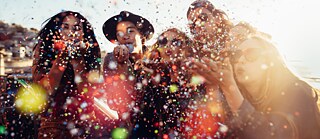 Skupina ľudí stojí blízko seba a fúka farebné konfety smerom ku kamere. Slnečné svetlo vytvára jasné odlesky a pestré svetelné body v zábere.