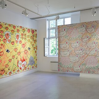 Ein heller Ausstellungsraum mit weißen Wänden zeigt zwei großformatige Wandarbeiten. Links hängt ein gelbes Werk mit dicht angeordneten orange‑roten Blütenformen, dazwischen stilisierte Tierfiguren. Rechts befindet sich ein großes Werk in Natur‑ und Pastelltönen, auf dem sich wiederholende, konturenhafte Darstellungen von Vogel‑ und Pflanzenmotiven angeordnet sind. Beide Arbeiten sind direkt auf die Wandfläche montiert. Die Fenster im Hintergrund lassen Tageslicht in den Raum, und an der Decke sind mehrere Strahler zur Beleuchtung installiert. Der Boden ist glatt und hell gehalten.