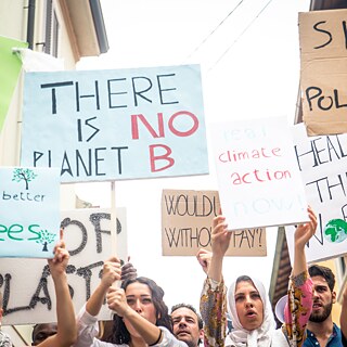 Eine Gruppe von Personen steht dicht beisammen in einer Straße und hält verschiedene Protestschilder in die Höhe. Die Schilder bestehen aus Karton- und Papierplatten in unterschiedlichen Größen und Farben. Darauf stehen Botschaften wie „THERE IS NO PLANET B“, „SICK OF POLLUTION“, „Life is better with trees“, „real climate action now“, „HEAL THE WORLD“ und weitere handgeschriebene Texte. Die Personen tragen unterschiedliche Alltagskleidung, und einige Arme sind deutlich erhoben, sodass die Schilder gut sichtbar sind. Die Straße ist schmal und von Gebäuden gesäumt, der Himmel ist hell.