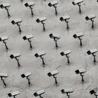 Eine große Anzahl identischer Überwachungskameras ist in gleichmäßigem Rastermuster an einer grauen Betonwand montiert. Jede Kamera ist auf einer kleinen Halterung befestigt und leicht schräg nach unten ausgerichtet. Die Kameras sind in diagonal versetzten Reihen angeordnet, sodass ein durchgängiges, sich wiederholendes Muster entsteht. Die Beleuchtung ist gleichmäßig und wirft weiche Schatten unter den Kameras.