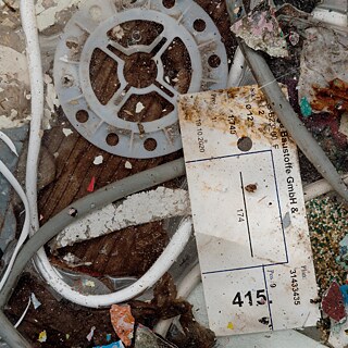 Ausschnitt einer Ansammlung von Bau- und Kunststoffabfällen, darunter Kabel, farbige Splitter, eine runde Kunststoffscheibe und ein verschmutztes beschriftetes Etikett.