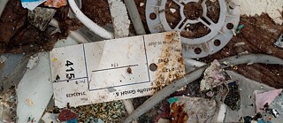 Ausschnitt einer Ansammlung von Bau- und Kunststoffabfällen, darunter Kabel, farbige Splitter, eine runde Kunststoffscheibe und ein verschmutztes beschriftetes Etikett.