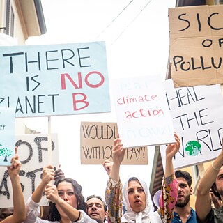 Eine Gruppe von Personen steht dicht beisammen in einer Straße und hält verschiedene Protestschilder in die Höhe. Die Schilder bestehen aus Karton- und Papierplatten in unterschiedlichen Größen und Farben. Darauf stehen Botschaften wie „THERE IS NO PLANET B“, „SICK OF POLLUTION“, „Life is better with trees“, „real climate action now“, „HEAL THE WORLD“ und weitere handgeschriebene Texte. Die Personen tragen unterschiedliche Alltagskleidung, und einige Arme sind deutlich erhoben, sodass die Schilder gut sichtbar sind. Die Straße ist schmal und von Gebäuden gesäumt, der Himmel ist hell.