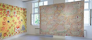 Ein heller Ausstellungsraum mit weißen Wänden zeigt zwei großformatige Wandarbeiten. Links hängt ein gelbes Werk mit dicht angeordneten orange‑roten Blütenformen, dazwischen stilisierte Tierfiguren. Rechts befindet sich ein großes Werk in Natur‑ und Pastelltönen, auf dem sich wiederholende, konturenhafte Darstellungen von Vogel‑ und Pflanzenmotiven angeordnet sind. Beide Arbeiten sind direkt auf die Wandfläche montiert. Die Fenster im Hintergrund lassen Tageslicht in den Raum, und an der Decke sind mehrere Strahler zur Beleuchtung installiert. Der Boden ist glatt und hell gehalten.