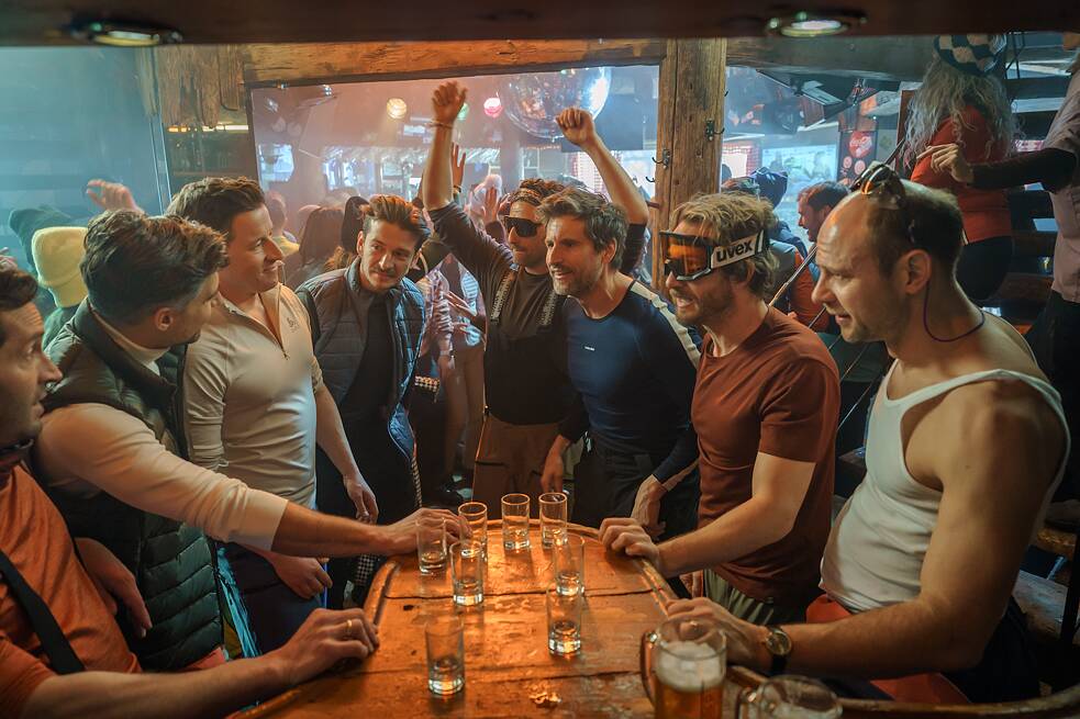 Still Frame from the Netflix Germany Series "Dudes": David Rott; Tom Beck; Moritz Führmann; Serkan Kaya in a bar setting drinking shots with another group of patrons standing around a table.