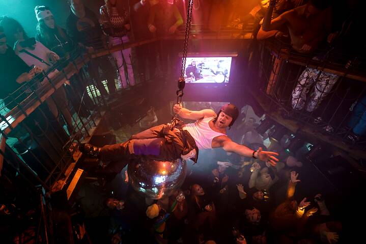 Still Frame from the Netflix Germany Series "Dudes", Moritz Führmann is riding a huge disco ball, dangling precariously over a party crowd.