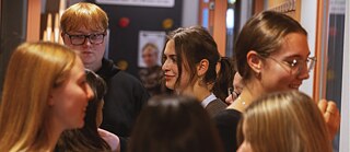 Eine Gruppe junger Menschen steht eng beieinander in einem Innenraum und unterhält sich. Die Umgebung wirkt lebendig und erinnert an eine Veranstaltung oder ein gemeinsames Treffen.