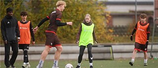 Zwei Jugendliche spielen Fußball auf einer Rasenfläche im Freien. Beide tragen Sportkleidung, während sie um den Ball kämpfen, umgeben von herbstlichen Bäumen und Trainingshütchen.