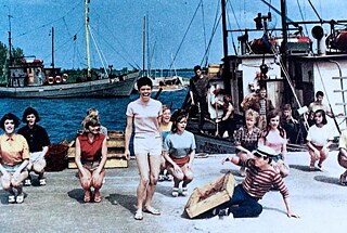 Das Standbild aus „Heißer Sommer / Hot Summer“ (DEFA 1968, directed by Joachim) zeigt eine Gruppe junger Menschen in farbenfroher Kleidung, die in einem Hafenszenario tanzt. „Hot Summer“ erlangte unter jungen Zuschauerinnen und Zuschauern in der DDR Kultstatus. Mit einer Formel aus Starappeal, populärer Musik und modisch gekleideten, attraktiven Jugendlichen orientierte sich der Film an westlichen Schlagerfilm‑Modellen.