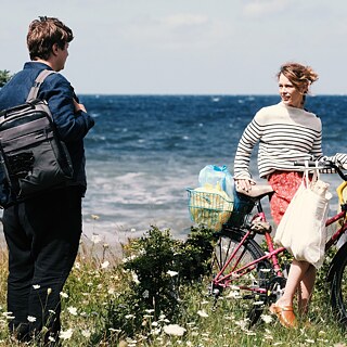 Dos personas están en una costa ventosa frente al mar. La mujer sujeta una bicicleta con bolsas de compra y una canasta con objetos, mientras el hombre está de pie frente a ella con una bolsa al hombro. Al fondo se observan las olas y el horizonte amplio.