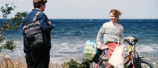 Dos personas están en una costa ventosa frente al mar. La mujer sujeta una bicicleta con bolsas de compra y una canasta con objetos, mientras el hombre está de pie frente a ella con una bolsa al hombro. Al fondo se observan las olas y el horizonte amplio.