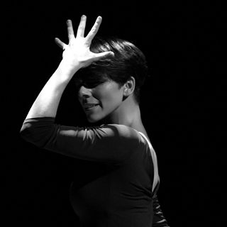 Schwarzer Hintergrund. Eine Frau hält ihre Hand mit gespreizten Fingern vor ihr Gesicht, der Kopf leicht gesenkt.