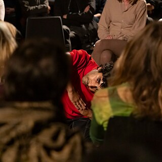 Eine Person in einem roten Oberteil führt eine performative Bewegung am Boden aus, neigt den Körper und blickt ins Publikum, umgeben von sitzenden Zuschauer:innen in einem schwach beleuchteten Raum.