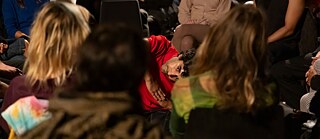 Eine Person in einem roten Oberteil führt eine performative Bewegung am Boden aus, neigt den Körper und blickt ins Publikum, umgeben von sitzenden Zuschauer:innen in einem schwach beleuchteten Raum.